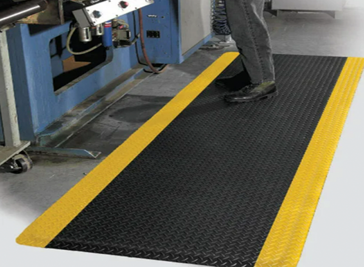 A person standing on a yellow and black mat in a factory, considering whether to buy anti-fatigue mats or hollow mats.